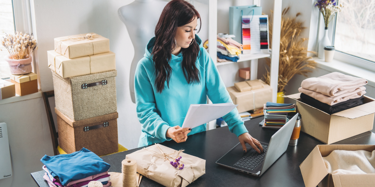 Small business owner working on a laptop beside packaged orders and clothing