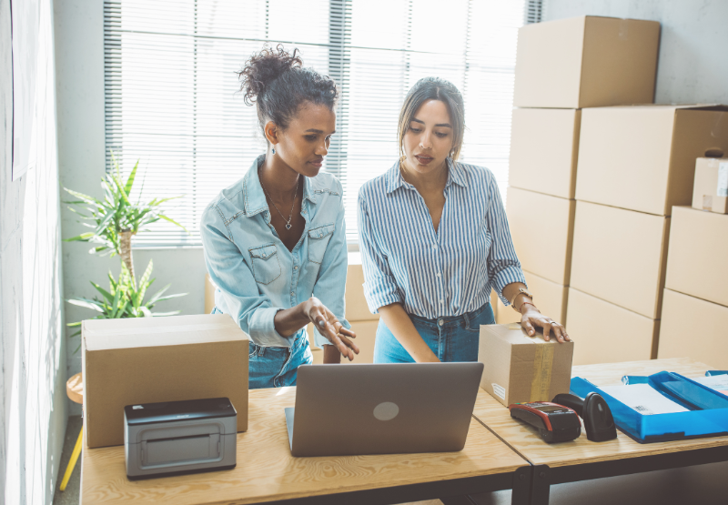 Deux personnes consultent des commandes sur un ordinateur dans une pièce remplie de boîtes d’expédition.