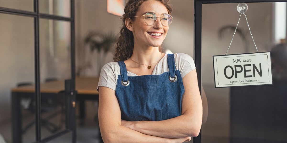 Small business owner standing beside an open sign at a storefront, representing business support and funding programs