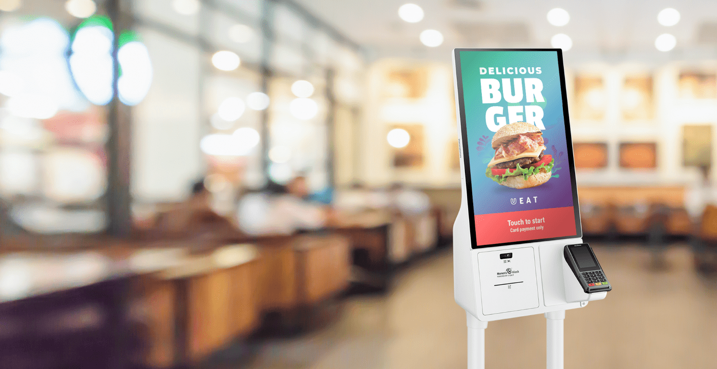 A Moneris Kiosk powered by UEAT on display in a quick-service restaurant with a delicious burger on the screen prompting the user to "order now."