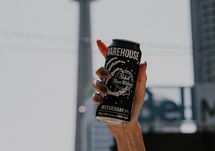 A hand holds a tall can of Warehouse branded Pabst Blue Ribbon beer