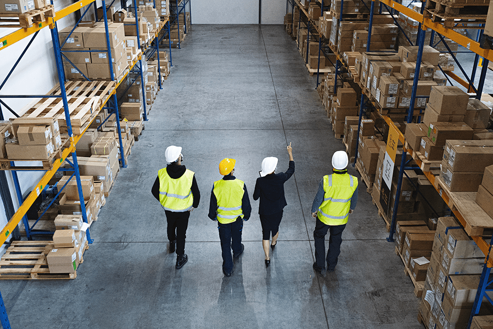 Four employees in a warehouse wearing yellow safety jackets walk down one of the aisles