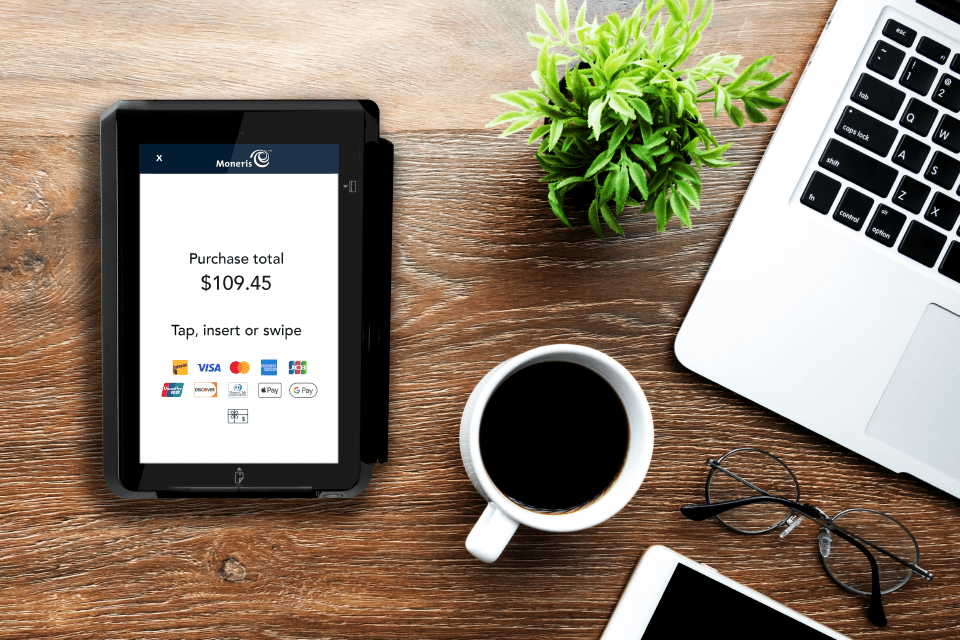 Moneris Go Unattended Device sits on a desktop beside a cup of coffee and an open laptop