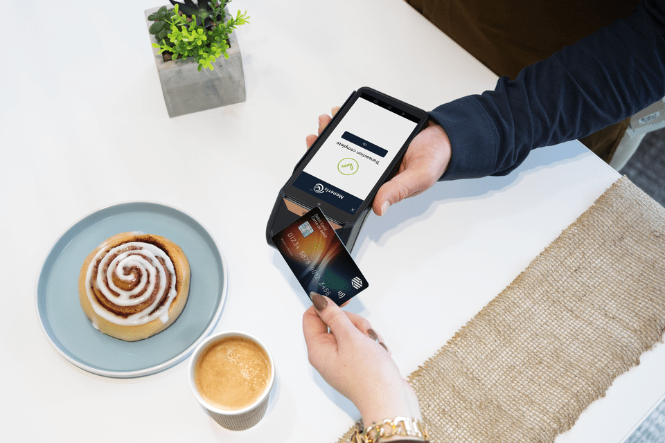 A customer taps a credit card on a Moneris Go Plus point-of-sale terminal held by a service provider, with a cinnamon roll and coffee on the table