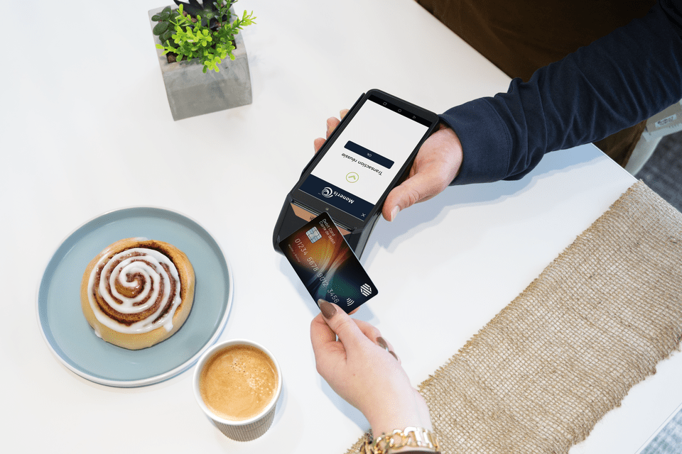 A customer taps a credit card on a Moneris Go Plus point-of-sale terminal held by a service provider, with a cinnamon roll and coffee on the table