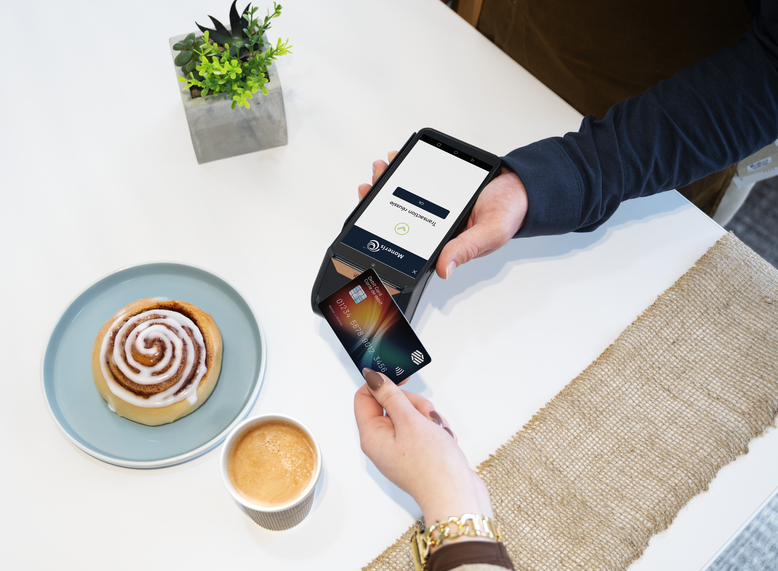 A customer taps a credit card on a Moneris point-of-sale terminal held by a service provider, with a cinnamon roll and coffee on the table
