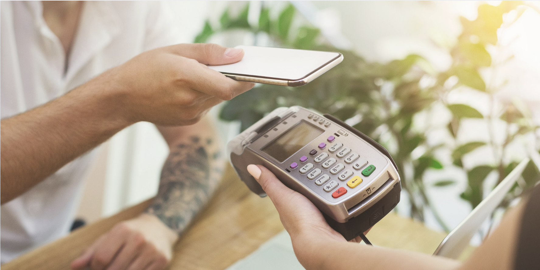 Customer taps a smartphone on a handheld payment terminal while a cashier holds the device at a café checkout.