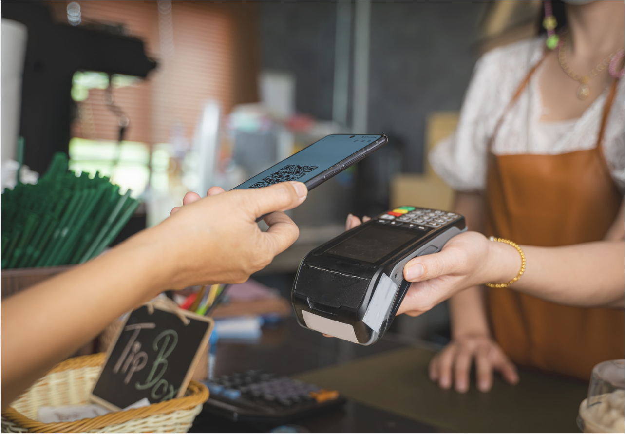 Customer taps a smartphone on a handheld payment terminal while a cashier holds the device at a café checkout.