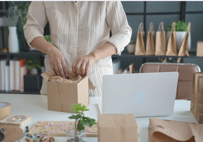 A small business owner carefully packages a customer order at a workstation with a laptop and shipping supplies, preparing products for delivery.