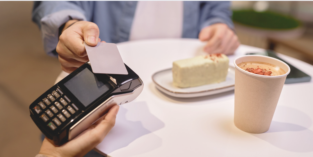 Customer taps a payment card on a handheld card terminal while seated at a café table with a coffee and slice of cake.