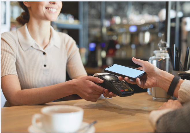 Customer holds a smartphone near a handheld payment terminal as a café employee accepts a contactless mobile payment at the counter.