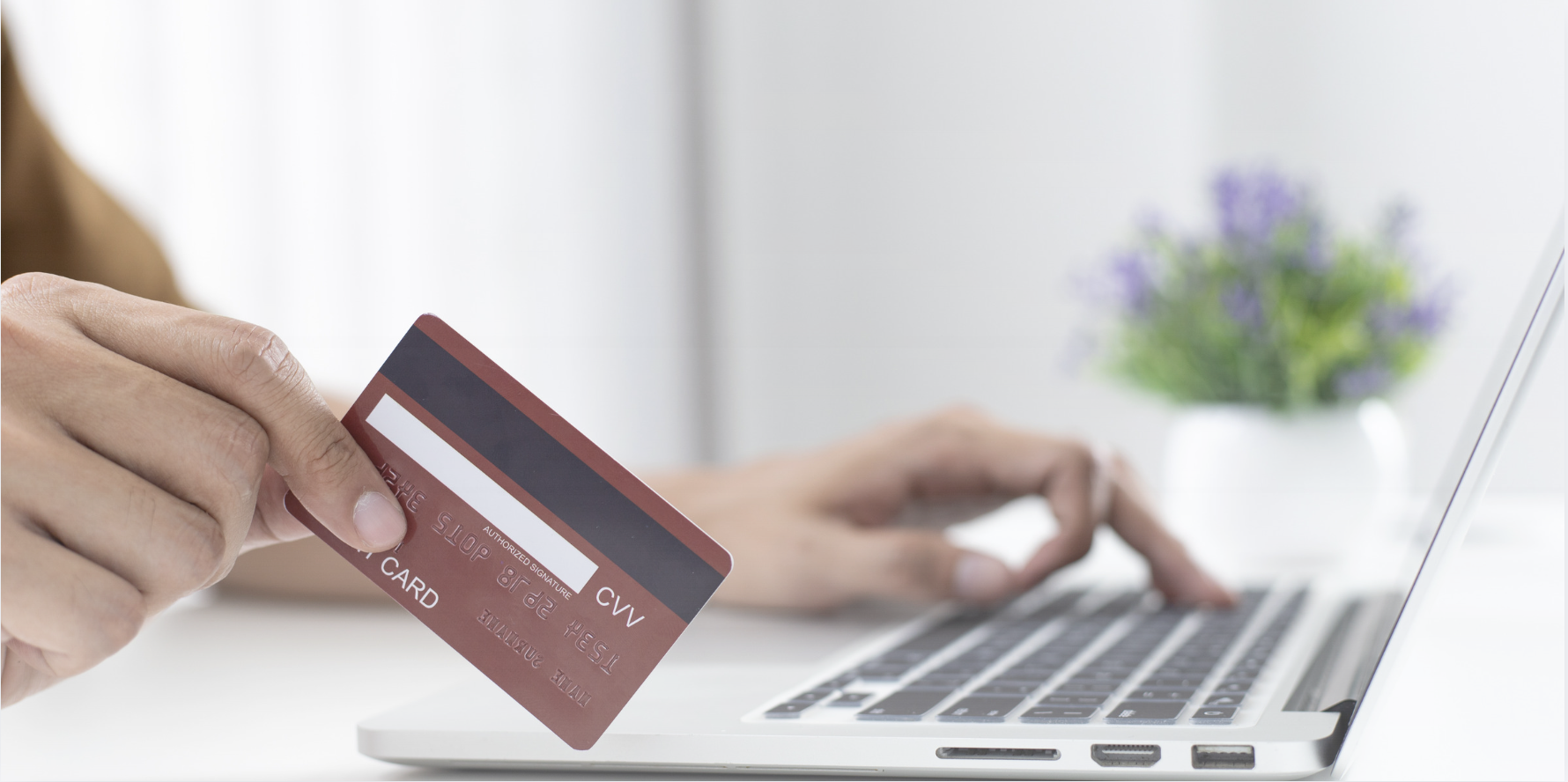 Person holding a credit card beside an open laptop while shopping online, with a coffee cup, smartphone, and eyeglasses on the desk in a home workspace.