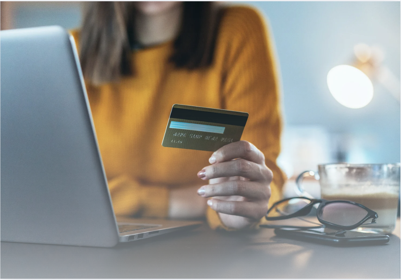 Person holding a credit card beside an open laptop while shopping online, with a coffee cup, smartphone, and eyeglasses on the desk in a home workspace.