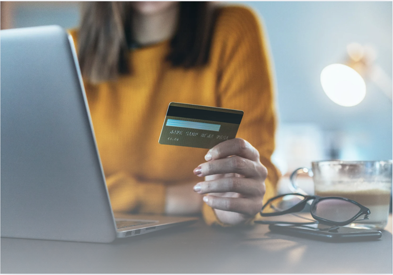 Person holding a credit card while using a laptop to complete an online purchase, with the card in focus and the computer keyboard blurred in the background.