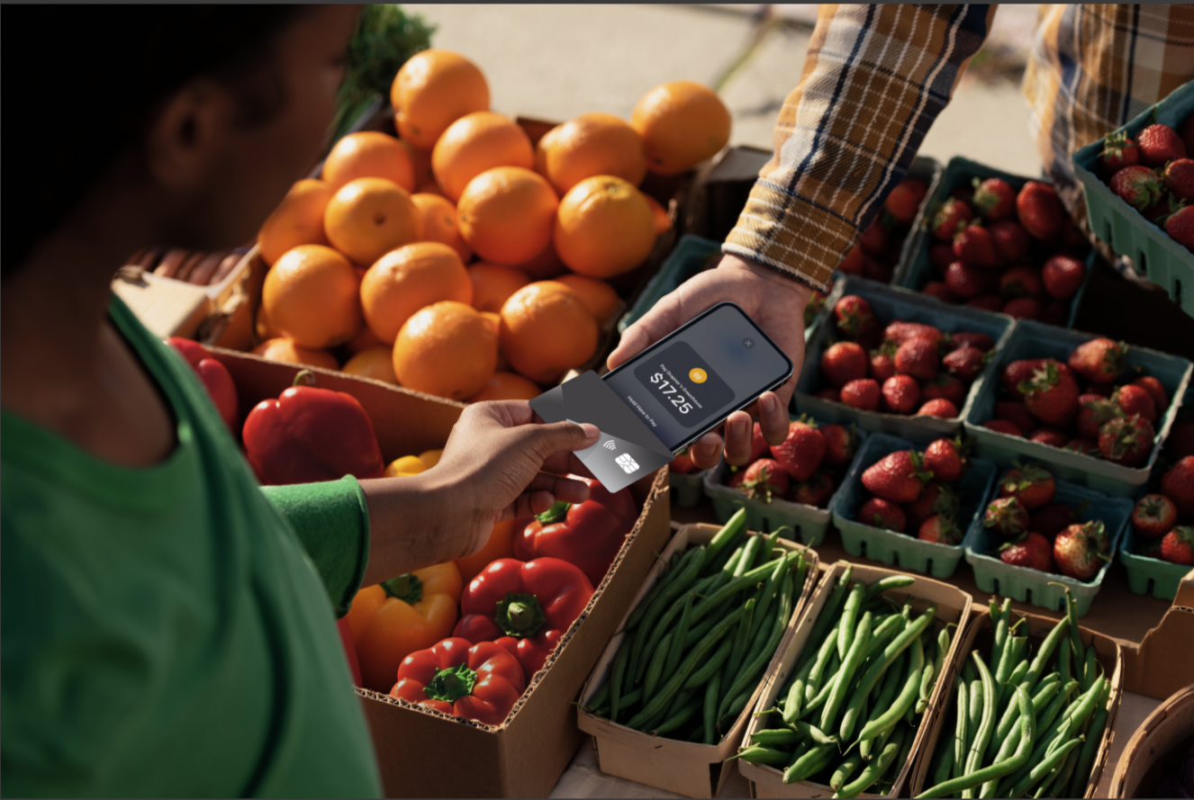 A customer at a farmers’ market holds a smartphone displaying a $17.25 payment amount while another person holds a payment card near the phone for a contactless transaction, surrounded by fresh oranges, strawberries, peppers, and green beans.
